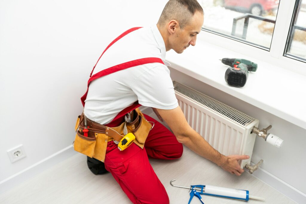 A male plumber installs a radiator in the heating system of an apartment. Guy in overalls and a gas